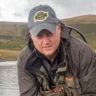 A man holding a small fish by a lake.