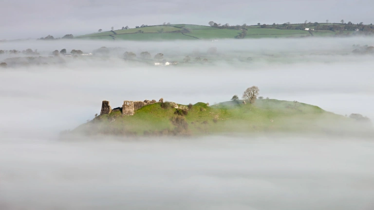 Golwg o bell o adfeilion castell ar ben bryn ynghanol cymylau isel 