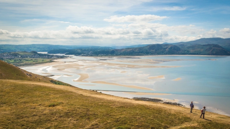 Dau berson yn syllu allan ar y môr â’r mynyddoedd yn y cefndir 