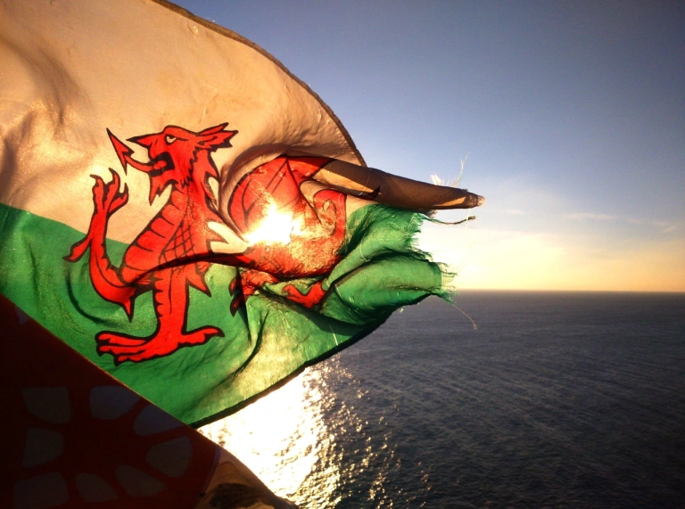 Baner Cymru yn fflachio yn erbyn y machlud gyda'r môr y tu ôl