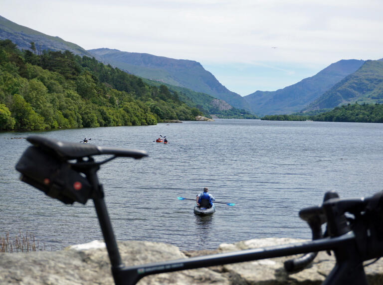 eic yn gorffwys ar wal gerrig, o flaen llyn eang gyda phobl yn canŵio ar y dŵr.