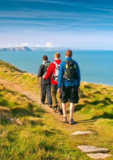 Tri dyn mewn dillad cerdded yn crwydro ar hyd Llwybr Arfordir Cymru gyda golygfeydd o’r môr 