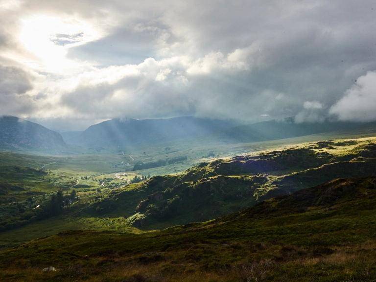 Pelydrau'r haul yn disgleirio trwy'r cymylau ar gaeau a bryniau