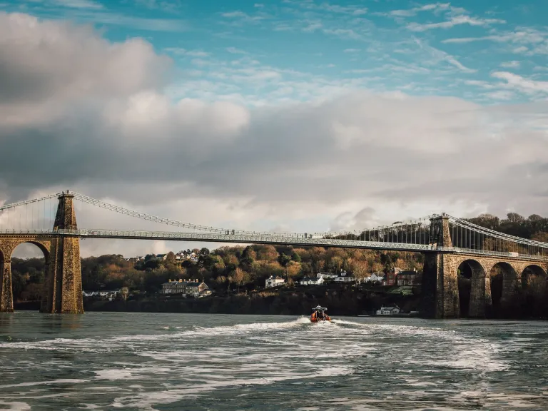 Llun o'r bont grog gyda choed a thai yn y cefndir a chwch ar Afon Menai