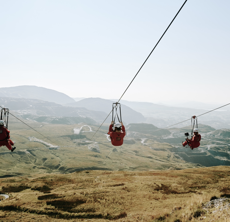 Tri o bobl yn marchogaeth zip-line yn uchel uwchben chwarel lechi.