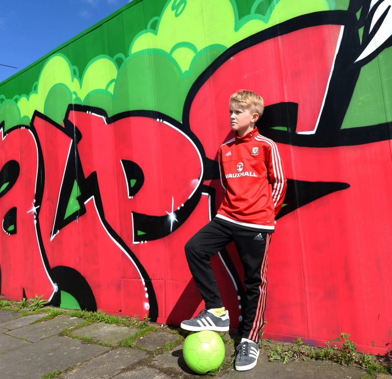 Bachgen yn pwyso ar wal gyda graffiti 'Cymru' arno, gydag un droed ar bêl-droed