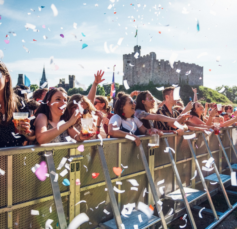 Cynulleidfa gig cerddoriaeth yn Tafwyl yn sefyll tu nôl i wahanfur gyda chonffeti o'u cwmpas a Chastell Caerdydd yn y cefndir