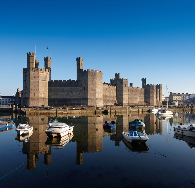 Llun o Gastell Caernarfon o'r de, gyda chychod yn yr harbwr o flaen y castell