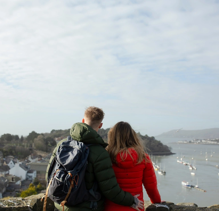 dyn a dynes yn sefyll mewn castell gyda golygfa o’r môr a chychod.