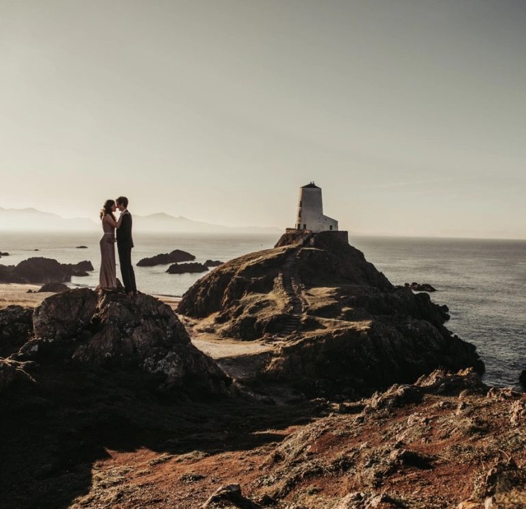 Llun priodas o gwpl yn cusanu ar ben craig ar draeth ynys Llanddwyn. Mae goleudy wen yn y cefndir.