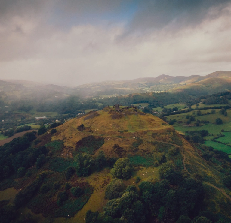 Llun o'r awyr o gastell ar fryn yng nghanol caeau gwyrdd. Mae'r awyr yn llwyd a chymylog. 